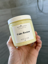 Hand holding a jar of 'Cake Batter' body scrub with a label on a neutral background