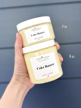 Hand holding two jars of 'Cake Batter' body scrub with visible labels.