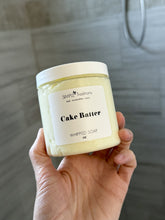 Hand holding a jar of 'Cake Batter' whipped soap with a label against a tiled wall background.