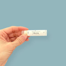 Hand holding a lip balm stick labeled 'Cherry' against a light blue background