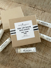 A set of Simple Traditions lip balms arranged on a rustic wooden surface, including a kraft paper box labeled 'THE LIP BALM SITUATION' and several white tubes with black text such as 'THE BACKUP', 'NIGHTSAND', and 'MANGROO BAG'.